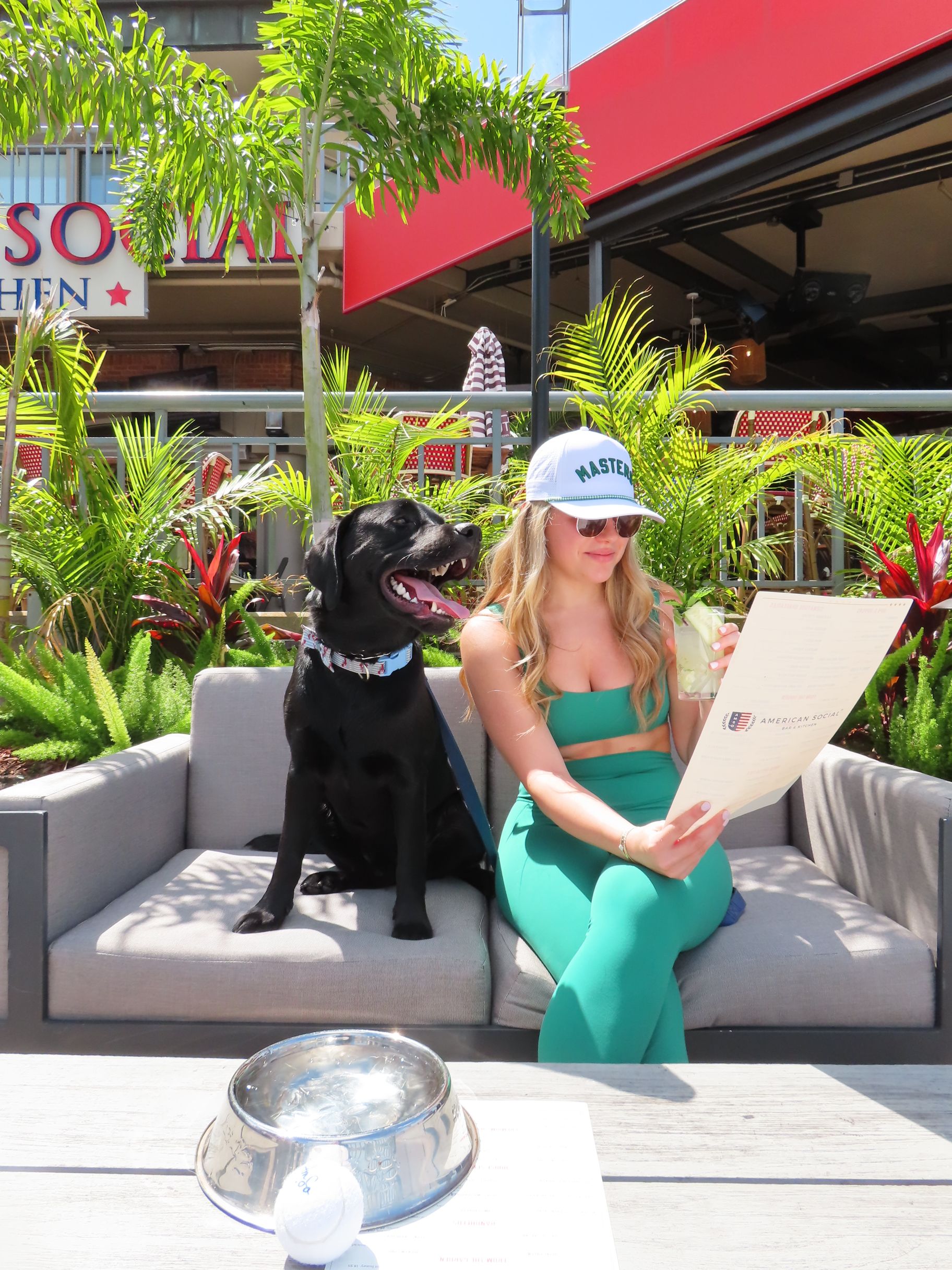 A woman in a green outfit and white cap sits on an outdoor couch with a black dog, reading a menu. They are surrounded by plants, and a shiny dog bowl is on the table in front of them.