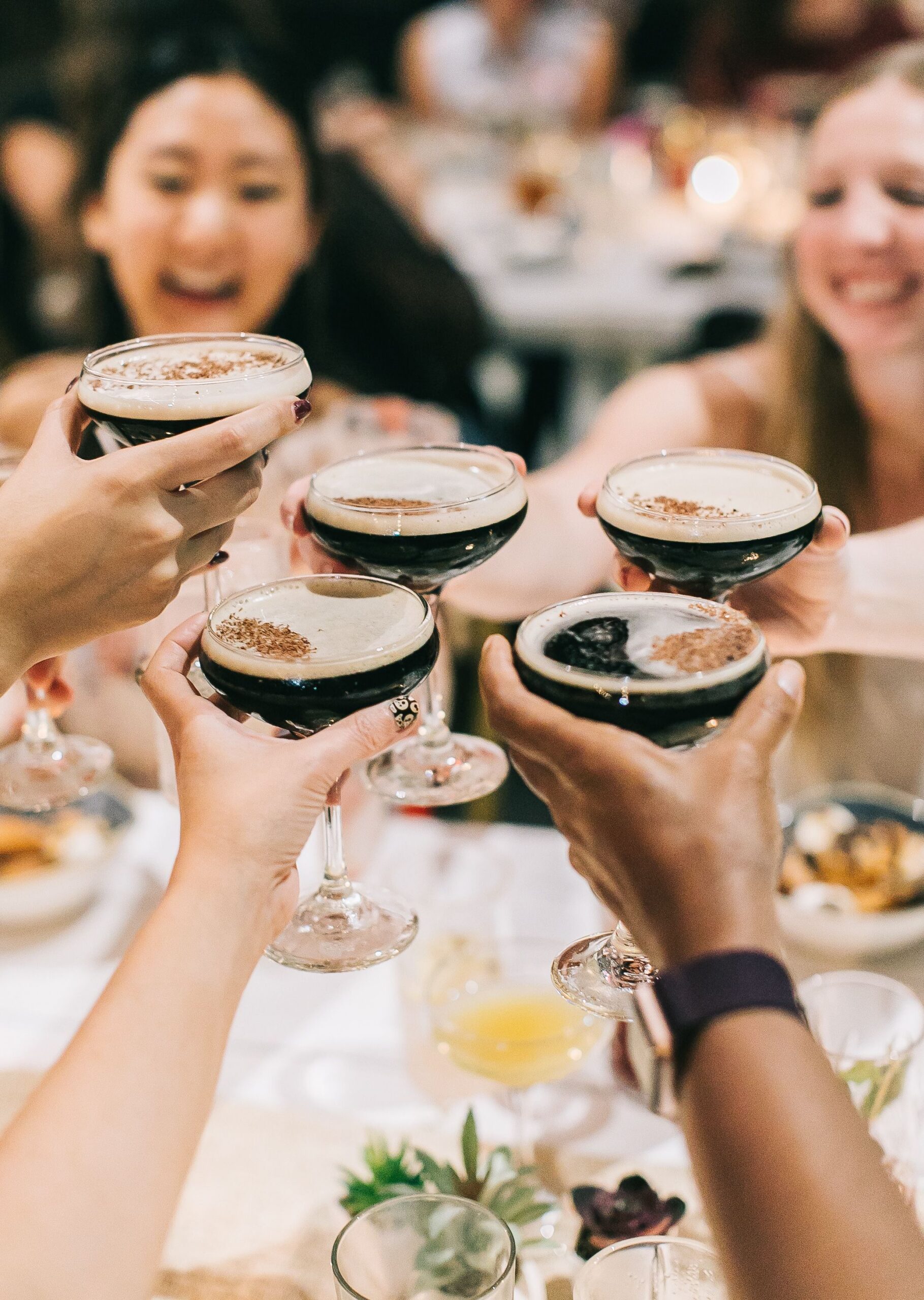 Five people clink glasses filled with a dark beverage at a lively event, smiling and celebrating around a table set with food.