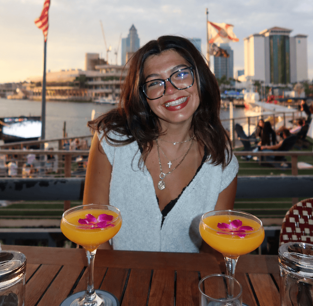 A woman with glasses smiles at a table with two yellow cocktails garnished with pink flowers. A waterfront cityscape and flags are visible in the background at sunset.