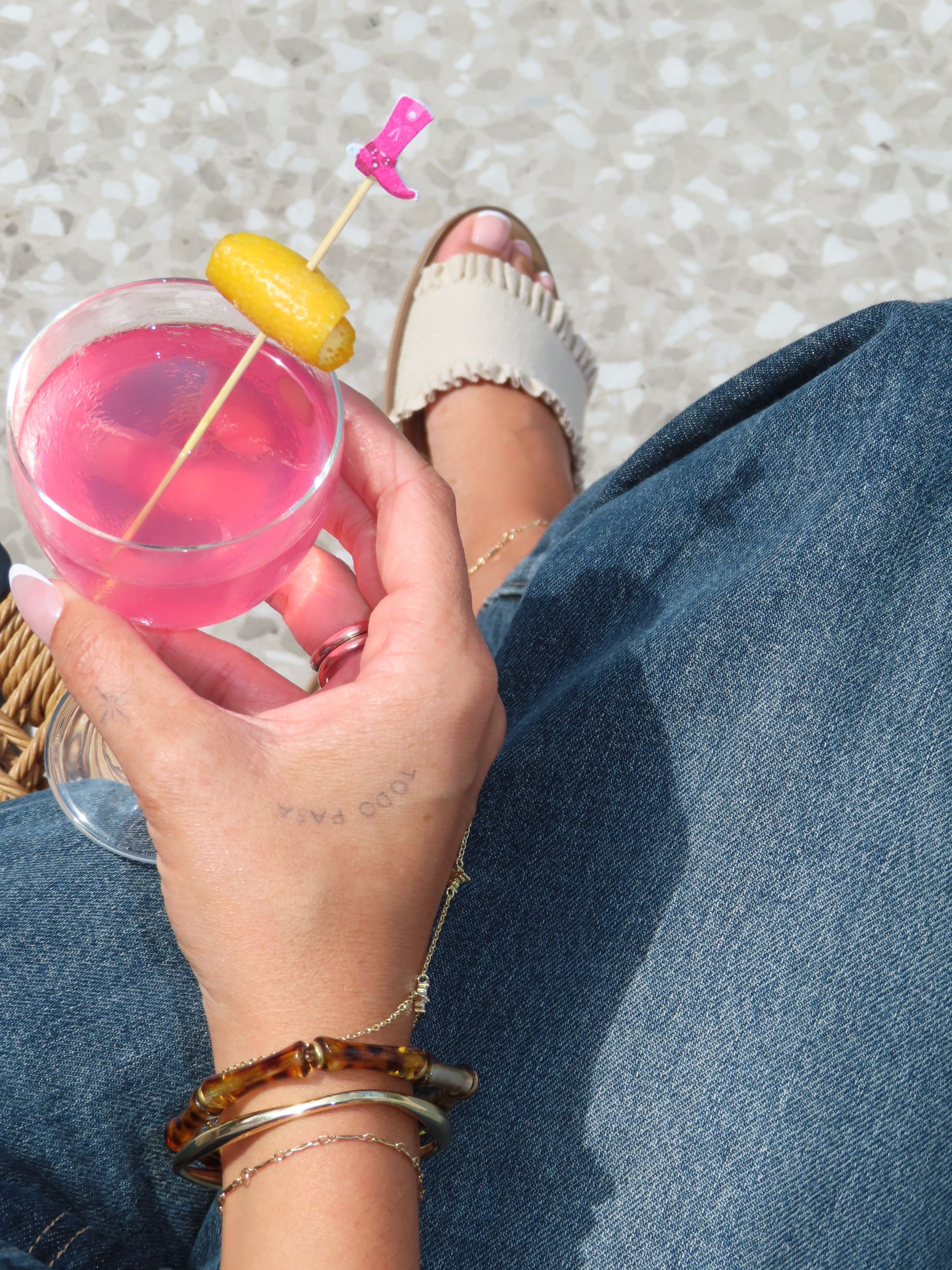 A hand holds a pink cocktail at an event, garnished with a lemon peel and a small pink cowboy boot pick.