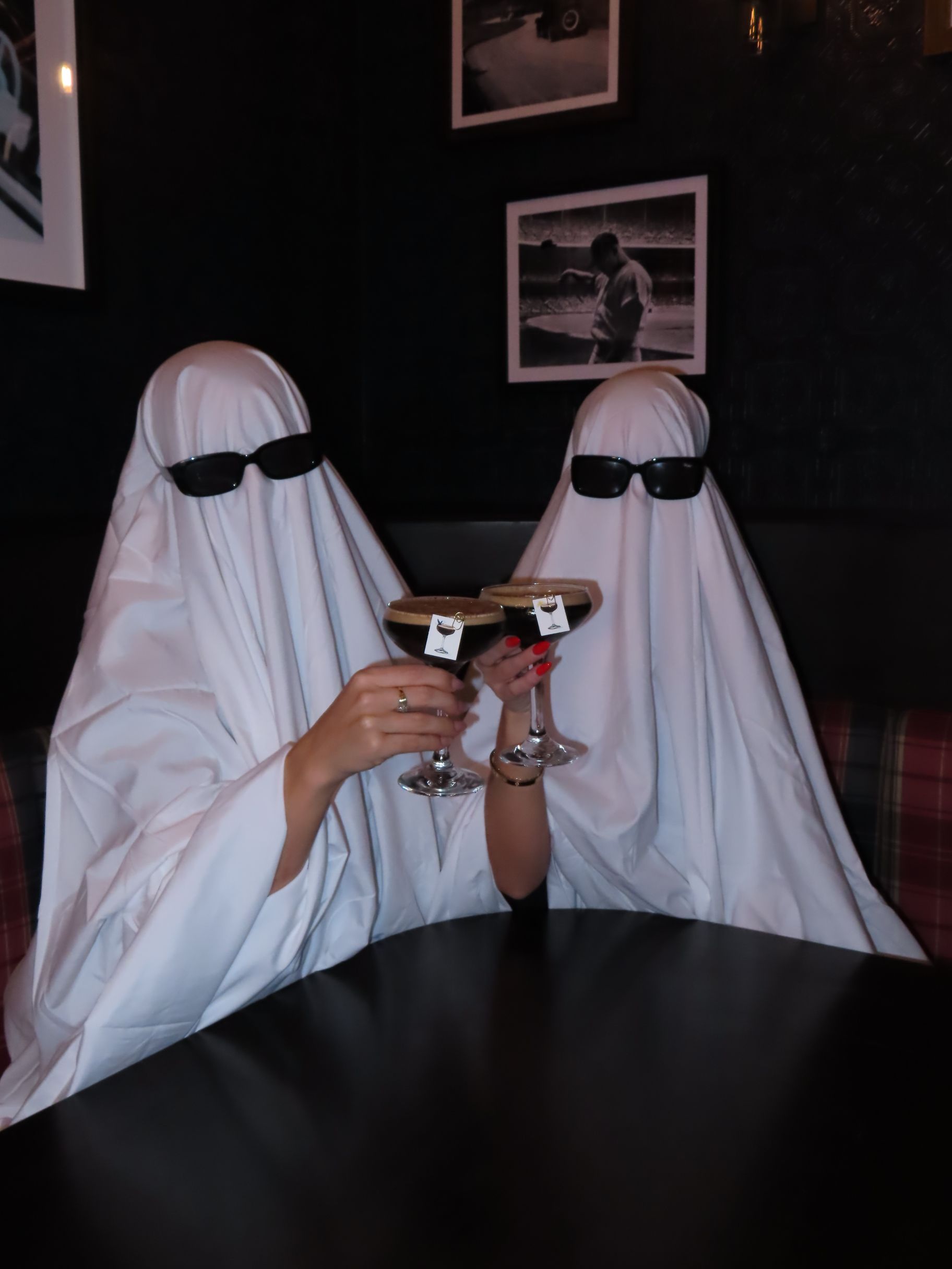 Two people dressed as ghosts wearing sunglasses toast with espresso martinis at an event table.