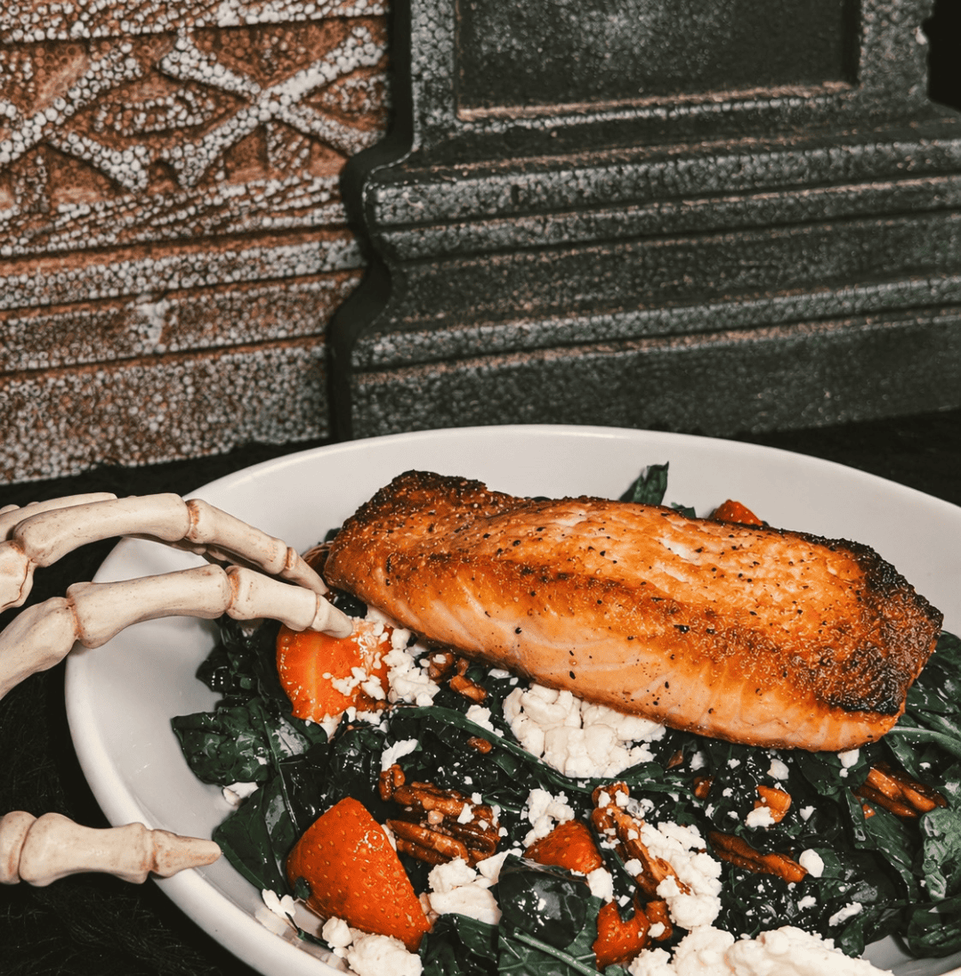 A plate of cooked salmon served over a salad of spinach, strawberries, pecans, and feta cheese, featuring decorative skeleton hands resting by a tombstone for a spooky themed event.