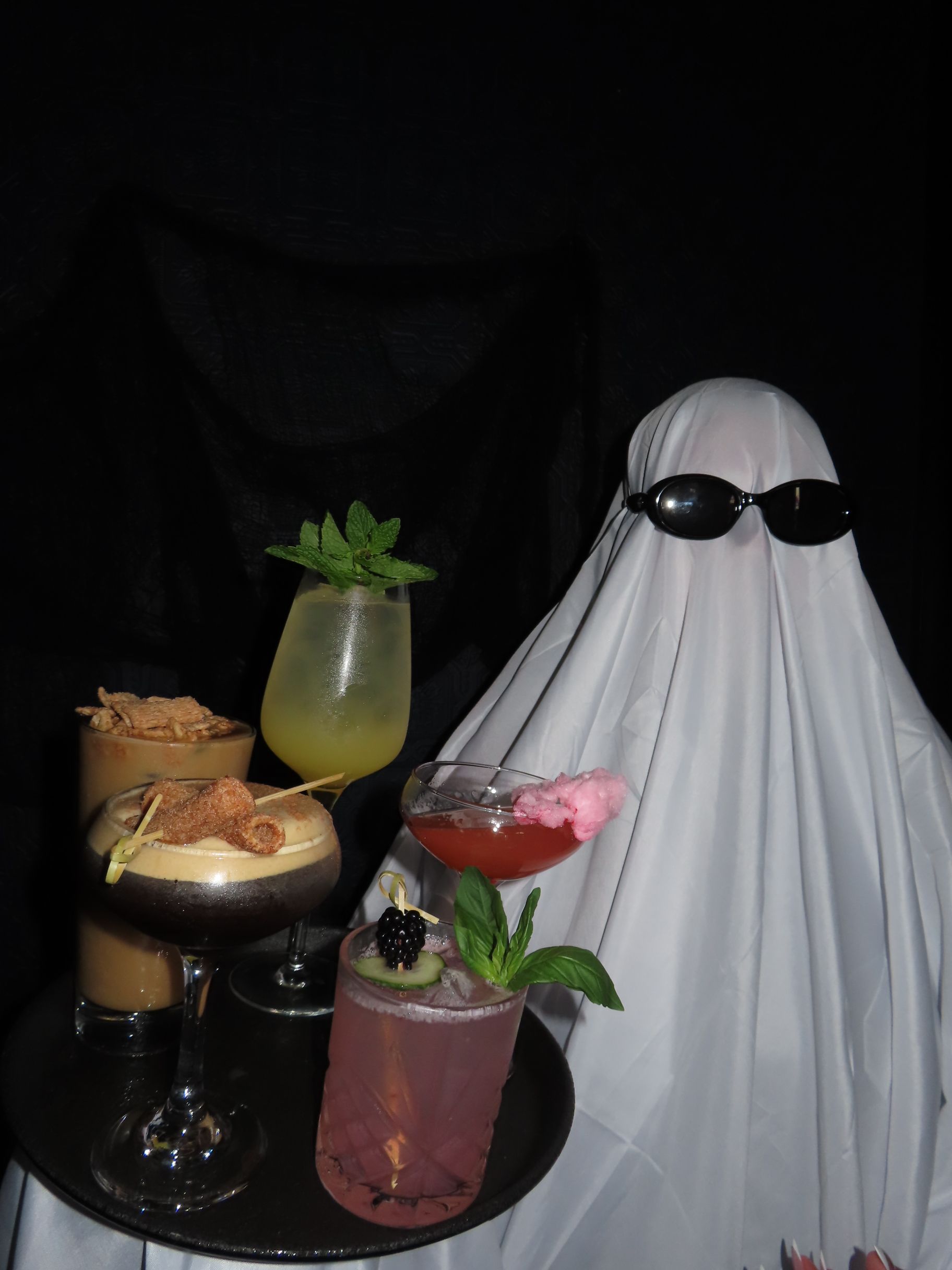 A person dressed as a ghost wearing sunglasses holds a tray of four various cocktails at an event.