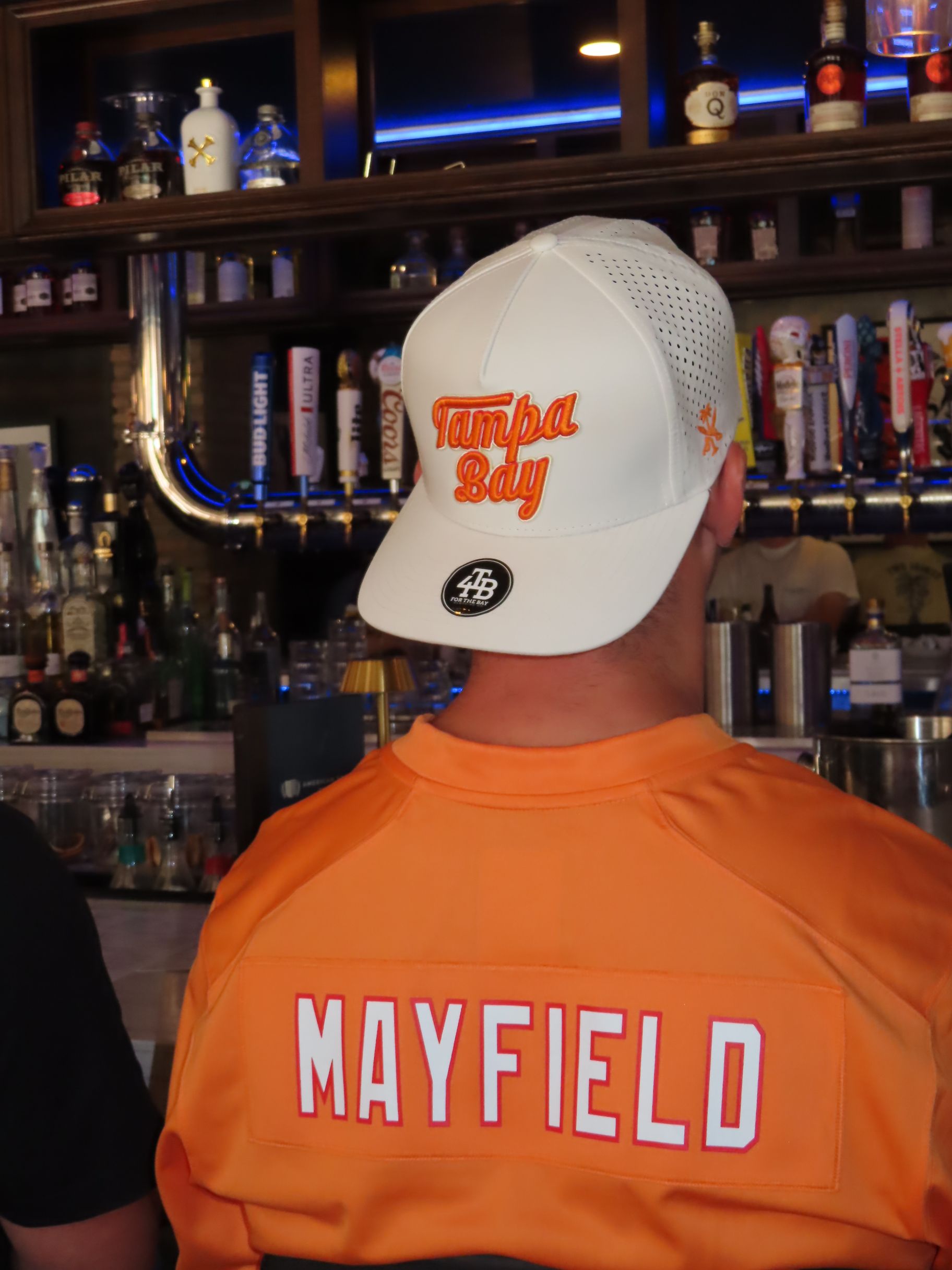 A person wearing an orange Mayfield jersey and a white Tampa Bay baseball cap stands in front of a bar during an event.